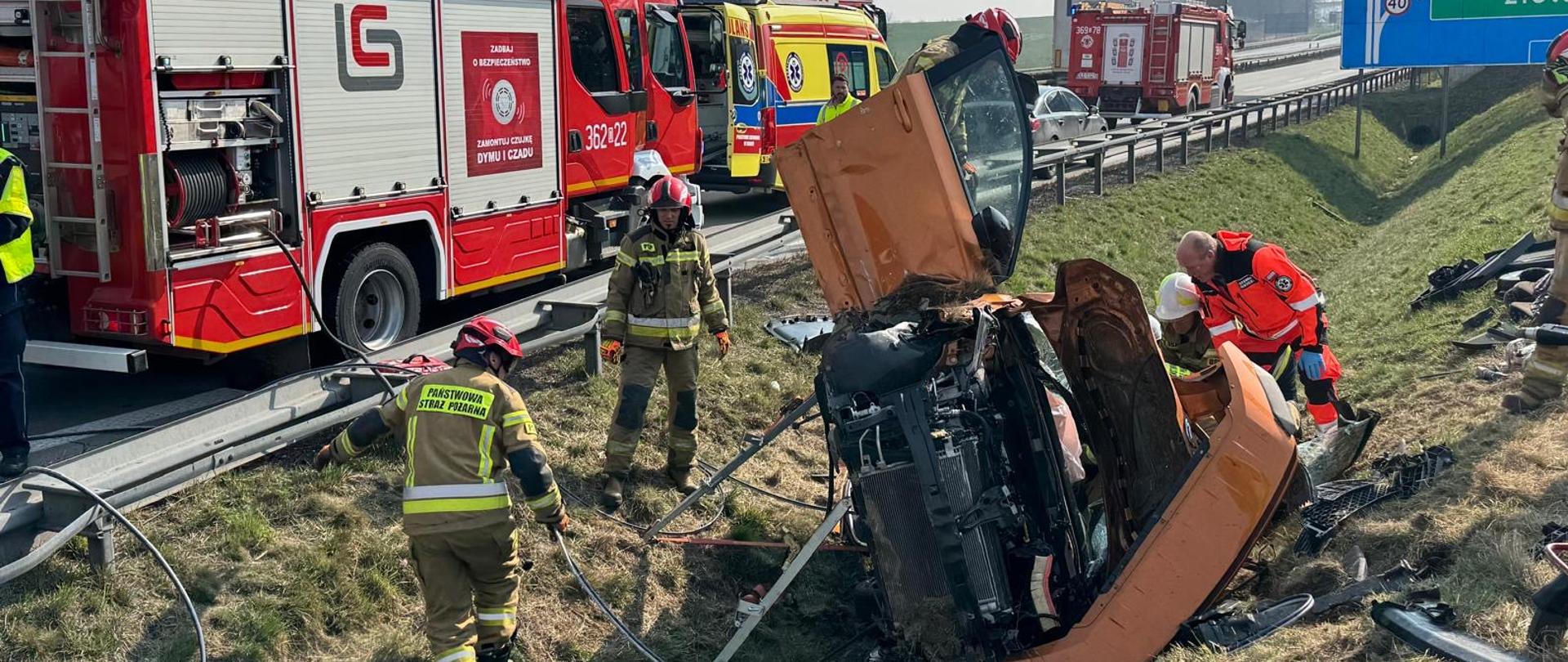 Zdjęcie przedstawia akcję ratunkową na autostradzie A4 w kierunku Wrocławia, po wypadku z udziałem pomarańczowego samochodu osobowego. Rozbity pojazd leży na boku w przydrożnym rowie, a ratownicy medyczni w pomarańczowych strojach oraz strażacy z Państwowej Straży Pożarnej pracują bezpośrednio przy wraku. W tle, na pasie awaryjnym, zaparkowane są pojazdy ratownicze: duży czerwony wóz strażacki o numerze operacyjnym 362[p]22, żółty ambulans oraz kolejny zastęp straży pożarnej w oddali. Nad drogą widoczna jest tablica informacyjna wskazująca kierunek na Wrocław, Legnicę i Złotoryję. Akcja toczy się w pełnym słońcu na trawiastym poboczu.