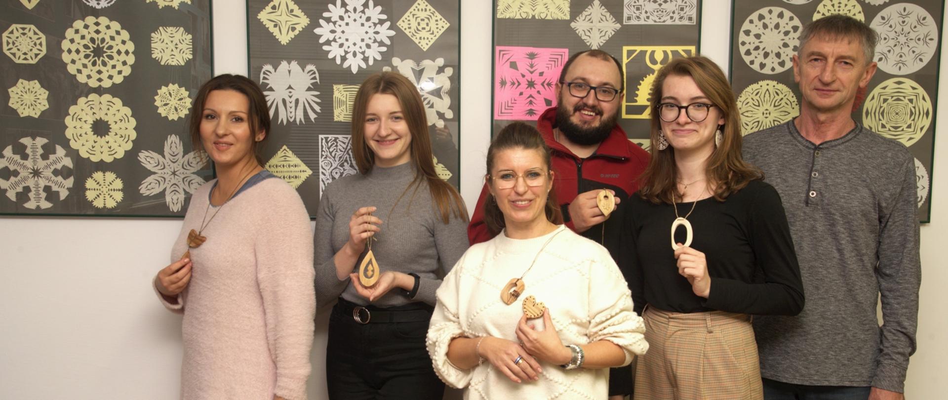 Na zdjęciu słuchacze oraz pan prowadzący warsztaty, stoją na tle wycinanek. Trzymają w rękach prace drewniane wykonane podczas warsztatów rękodzielniczych.