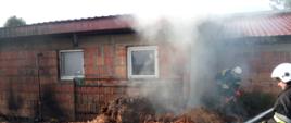 Murowany budynek inwentarski kryty blachą. Nad budynkiem widoczny gęsty dym. Przed budynkiem strażacy dogaszają wyniesione z budynku palące się resztki słomy.