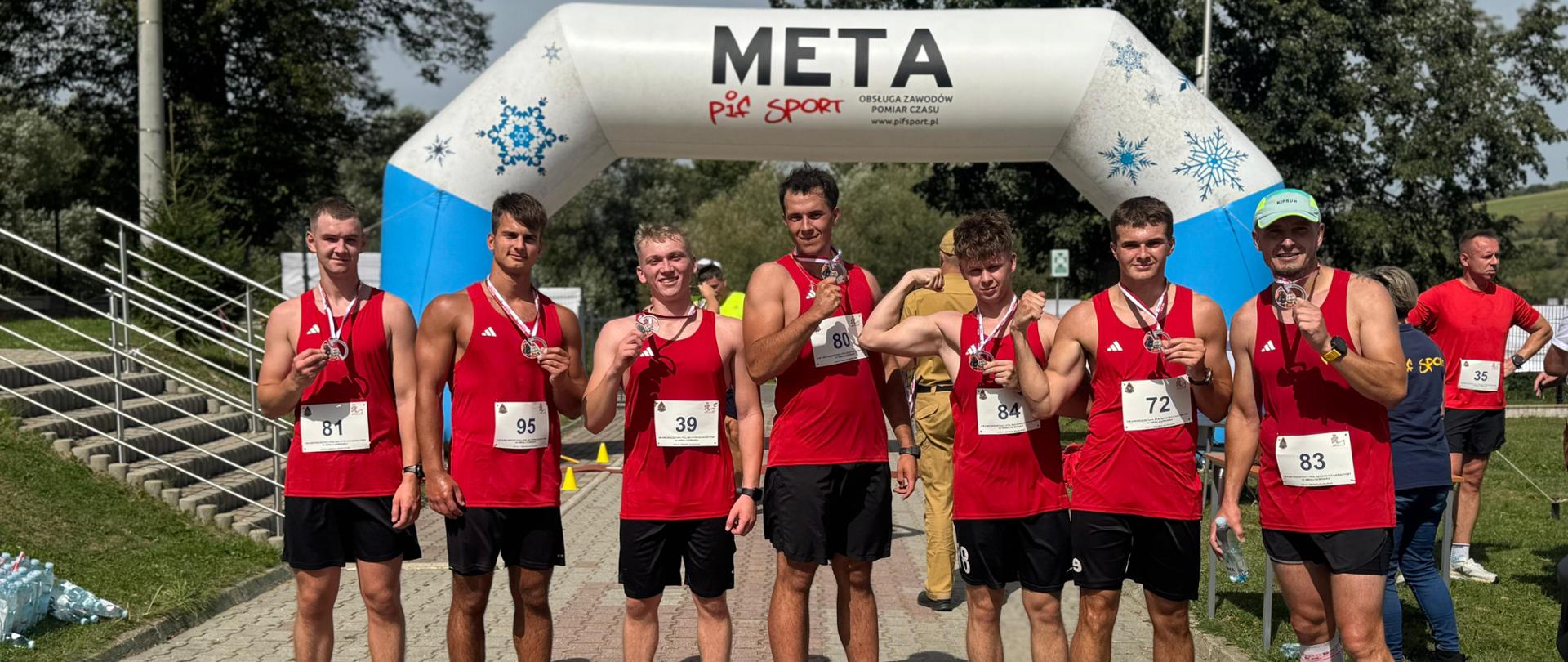 1. Na zdjęciu widać siedmiu młodych mężczyzn w czerwonych sportowych koszulkach i czarnych spodenkach, stojących w rzędzie pod dużym biało-niebieskim łukiem z napisem „META”. Każdy z nich ma przypięty numer startowy i trzyma w dłoni medal, który z dumą prezentuje do kamery. Zawodnicy uśmiechają się, część pokazuje zaciśnięte pięści w geście zwycięstwa. W tle widać innych uczestników zawodów, drzewa oraz słoneczną pogodę.