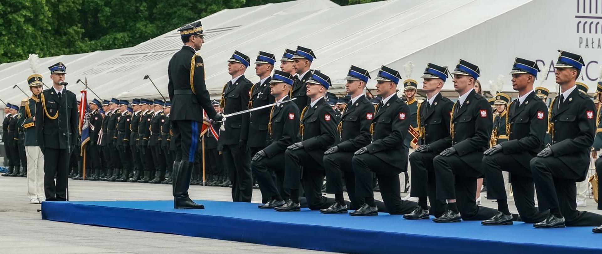 Strażaków, którzy klęczą na podeście i czekają na mianowanie na pierwszy stopień oficerski. Przed jednym z nich stoi Komendant Główny, który wykonuje ruch szablą na lewe ramię strażaka. 