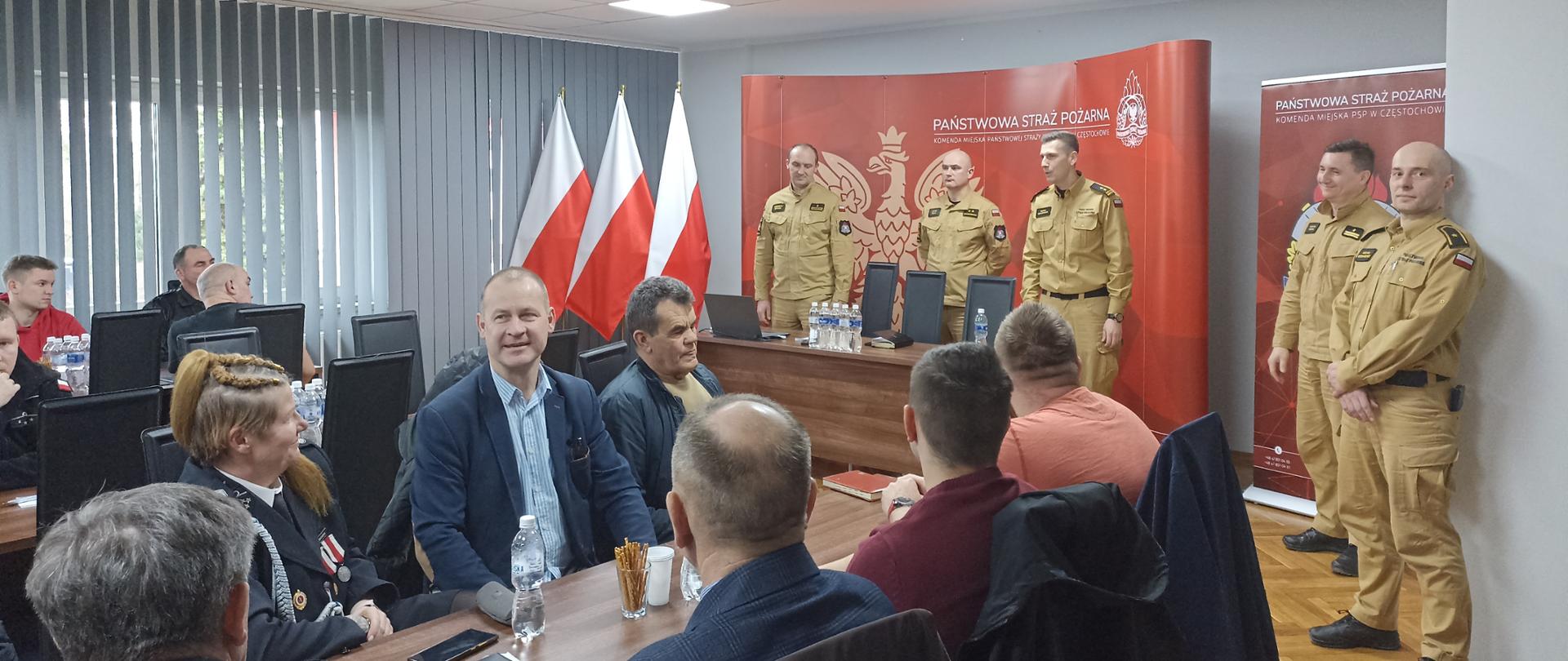 Na zdjęciu na sali przy stołach siedzą ludzie, przed nimi stoi pięciu funkcjonariuszy Państwowej straży pożarnej w umundurowaniu służbowym