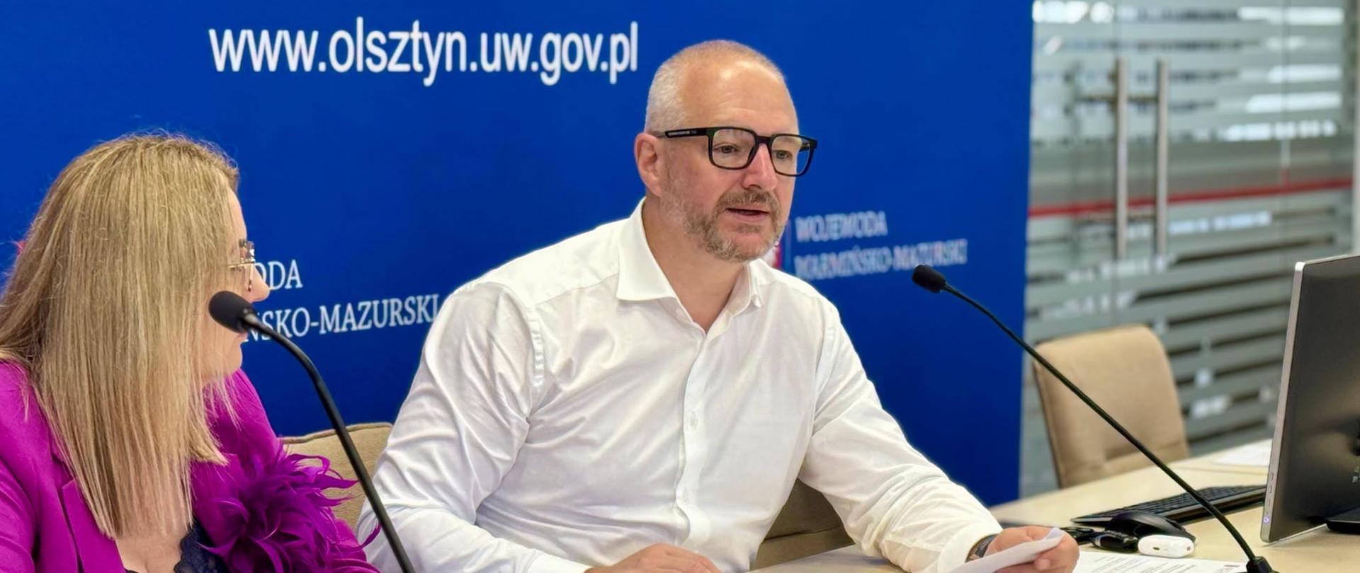 Mężczyzna w białej koszuli, wojewoda, przemawia przez mikrofon na tle niebieskiej ścianki 