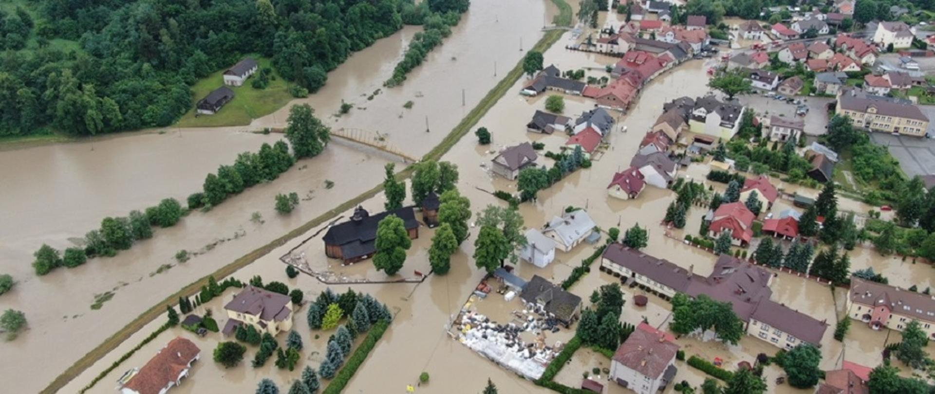 Zdjęcie przedstawia widok miejscowości pokazany z góry po powodzi. Widać budynki, zalane wodą posesje i ulice