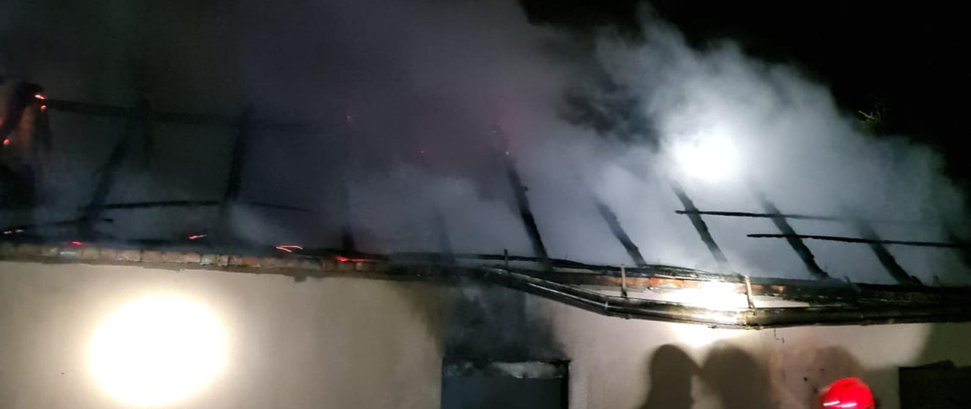 Widać budynek gospodarczy, spalony dach, z konstrukcji dachu wydobywa się dym, jest noc