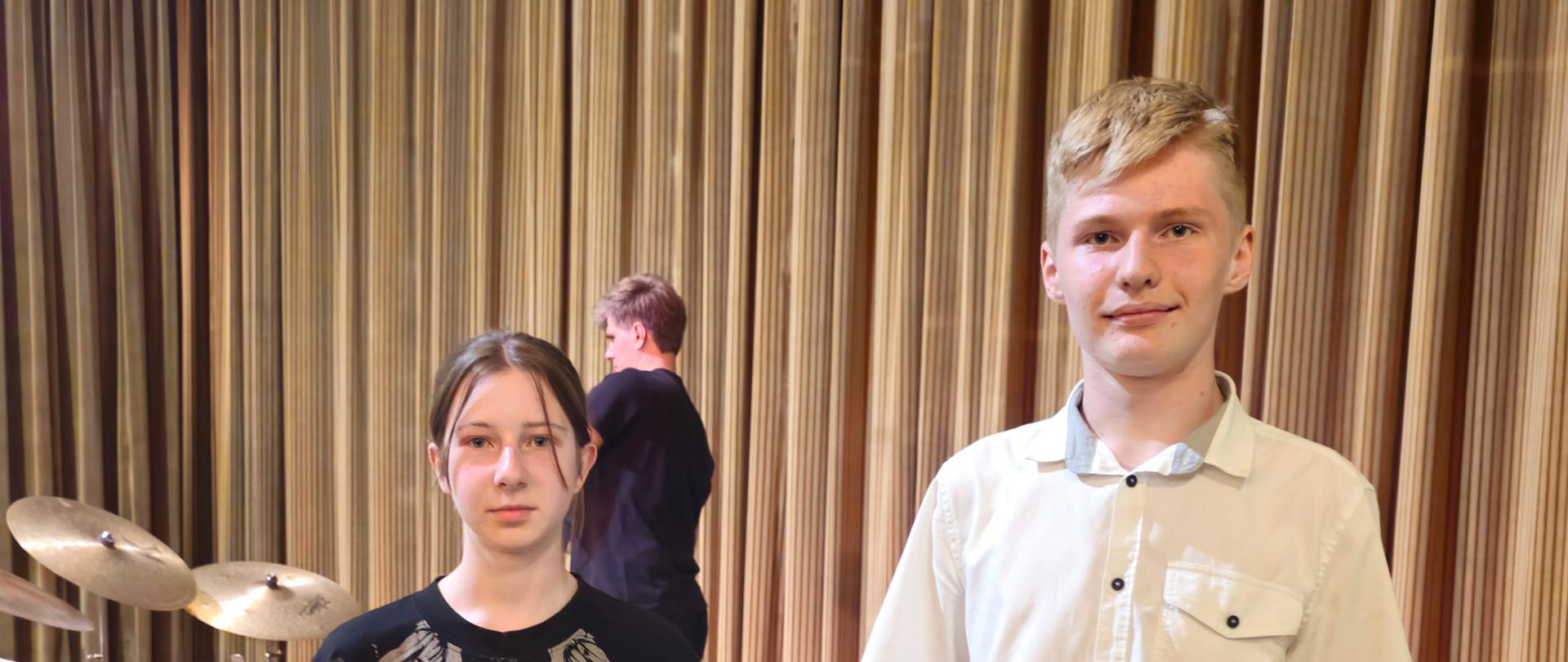 Fotografia przedstawia dwoje nastolatków, dziewczynę i chłopaka trzymających w ręku dyplomy, stojących obok siebie na podwyższonej drewnianej scenie w auli koncertowej. Tło stanowi brązowa zasłona przypominająca kurtynę, oraz zestaw perkusyjny widoczny w głębi po lewej stronie. W centralnej części tła, nieco z tyłu, widać trzecią osobę w czarnym ubraniu, odwróconą tyłem.