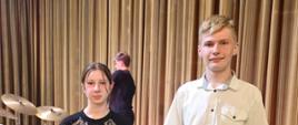 Fotografia przedstawia dwoje nastolatków, dziewczynę i chłopaka. Tło stanowi brązowa zasłona przypominająca kurtynę, oraz zestaw perkusyjny widoczny w głębi po lewej stronie. W centralnej części tła, nieco z tyłu, widać trzecią osobę w czarnym ubraniu, odwróconą tyłem.