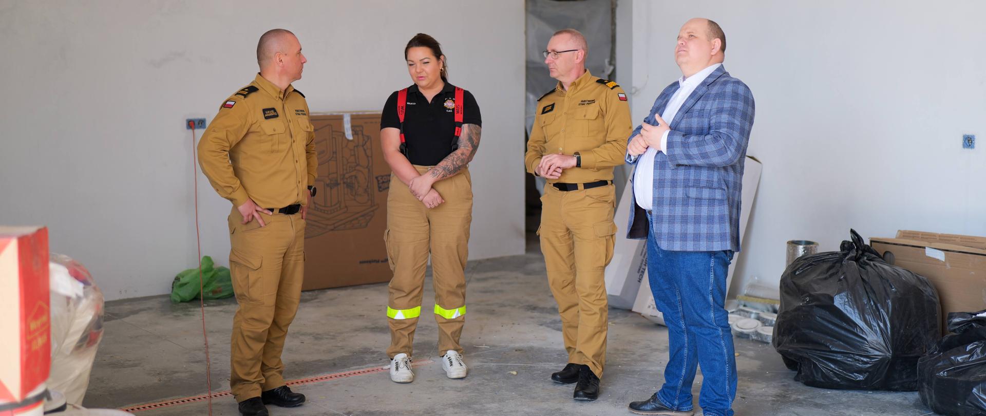 Cztery osoby – trzy w służbowych mundurach PSP i OSP oraz jedna w cywilnym ubraniu – stoją wewnątrz nowo budowanego budynku. Wokół widać materiały budowlane, kartony i worki. Osoby prowadzą rozmowę, przyglądając się wnętrzu obiektu.
