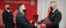 Na zdjęciu widocznych trzech strażaków PSP w mundurach galowych z założonymi maseczkami na twarzach podczas uroczystości w budynku KW PSP w Opolu.
Strażak po lewej stronie kadru - st. bryg. Krzysztof Kędryk Opolski Komendanta Wojewódzki PSP w Opolu wręcza upominek st. kpt. Sebastianowi Jantosowi Zastępcy Komendanta Powiatowego PSP w Kluczborku.