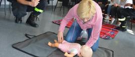 Szkolenie z pierwszej pomocy. Świetlica Komendy Powiatowej Policji w Rawiczu. Na pierwszym planie jedna z uczestniczek szkolenia, która klęcząc - pochyla się nad leżącym na kocu manekinie "niemowlaku" - wykonując czynności ratujące życie po uprzednim zakrztuszeniu się dziecka. 