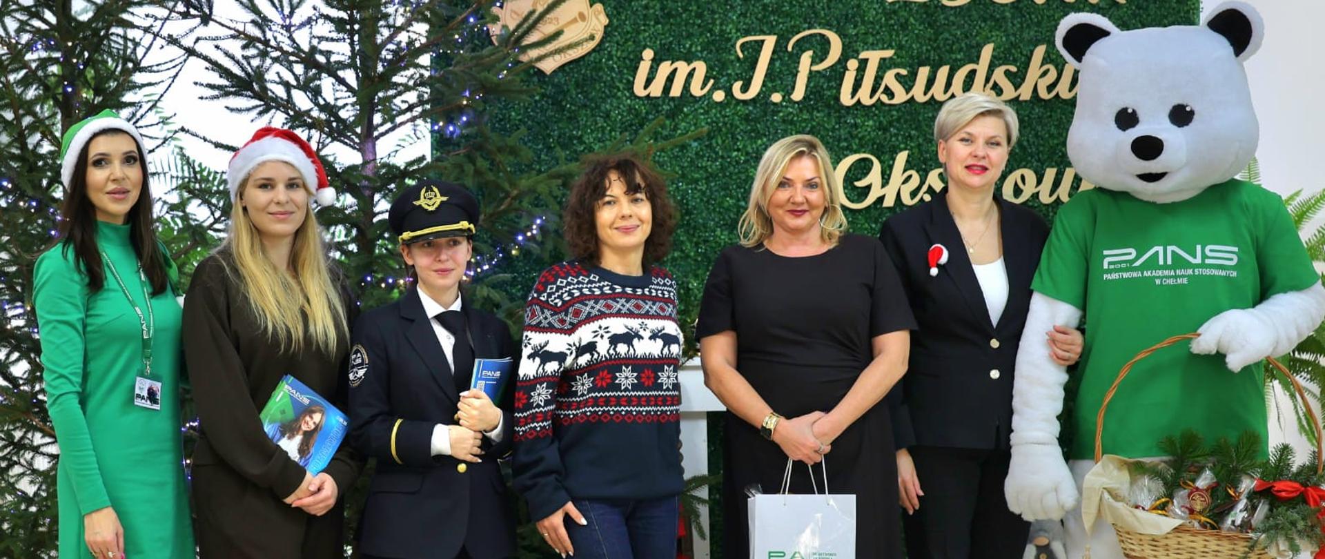 Na zdjęciu znajduje się grupa osób w świątecznych strojach, w tym pilotka i maskotka białego misia, stojąca obok choinek.