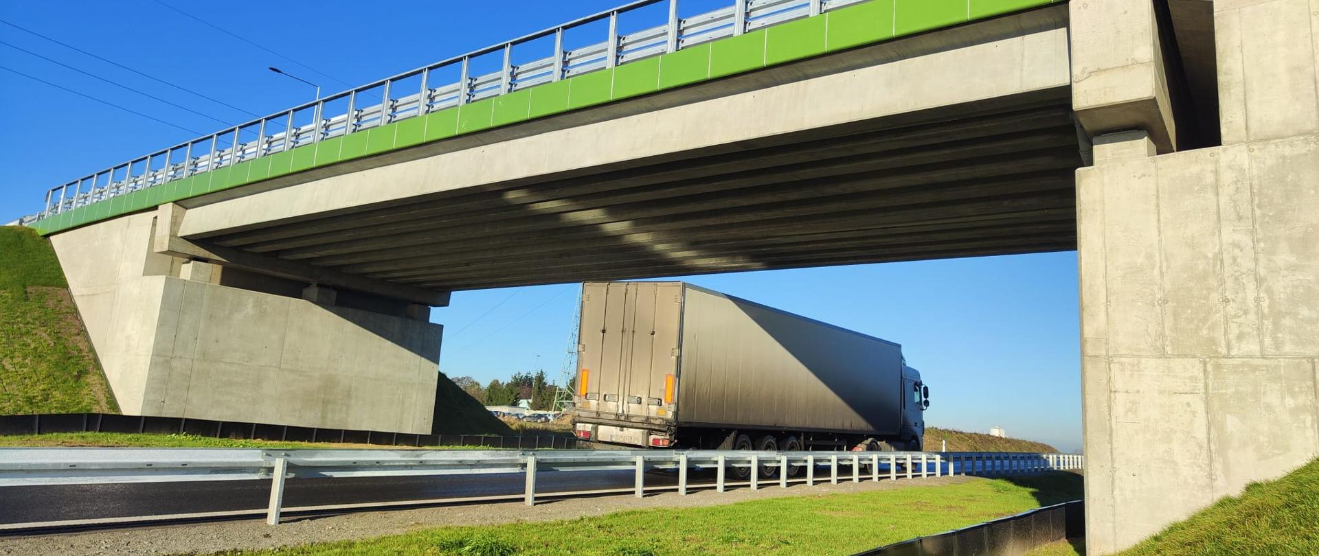 Zdjęcie przedstawia widok z poziomu drogi na wiadukt z zielonymi elementami wykończenia przy barierkach. Pod wiaduktem przejeżdża duży samochód ciężarowy z naczepą typu plandeka. 
