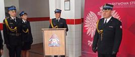 Nowo mianowany Dowódca Jednostki Ratowniczo Gaśniczej w Rawiczu przemawia do zgromadzonych na uroczystości strażaków oraz pracowników cywilnych Komendy. Po prawej stronie Komendant Powiatowy PSP w Rawiczu. Z lewej strony dowódca uroczystości oraz funkcjonariuszka odczytująca decyzję. W tle czerwona ścianka przenośna z godłem Polski.