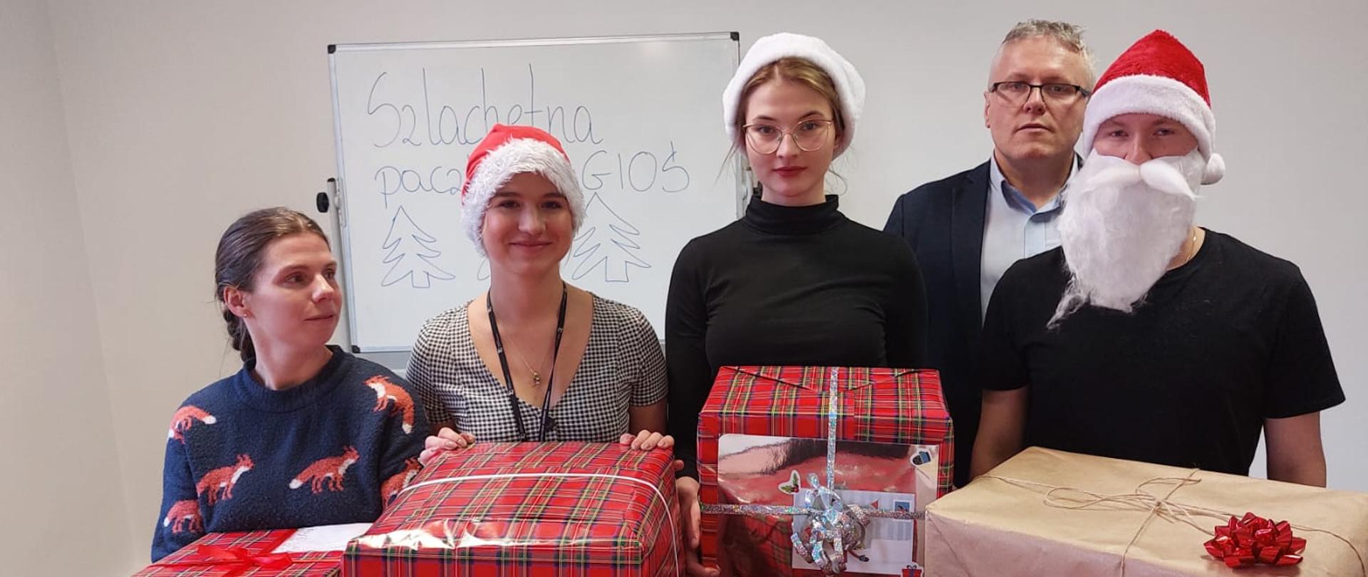 Stos paczek zapakowanych w czerwony papier . Z tyłu stoi i pozuje piątka osób, 3 kobiety i 2 mężczyzn.