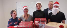 Stos paczek zapakowanych w czerwony papier . Z tyłu stoi i pozuje piątka osób, 3 kobiety i 2 mężczyzn.