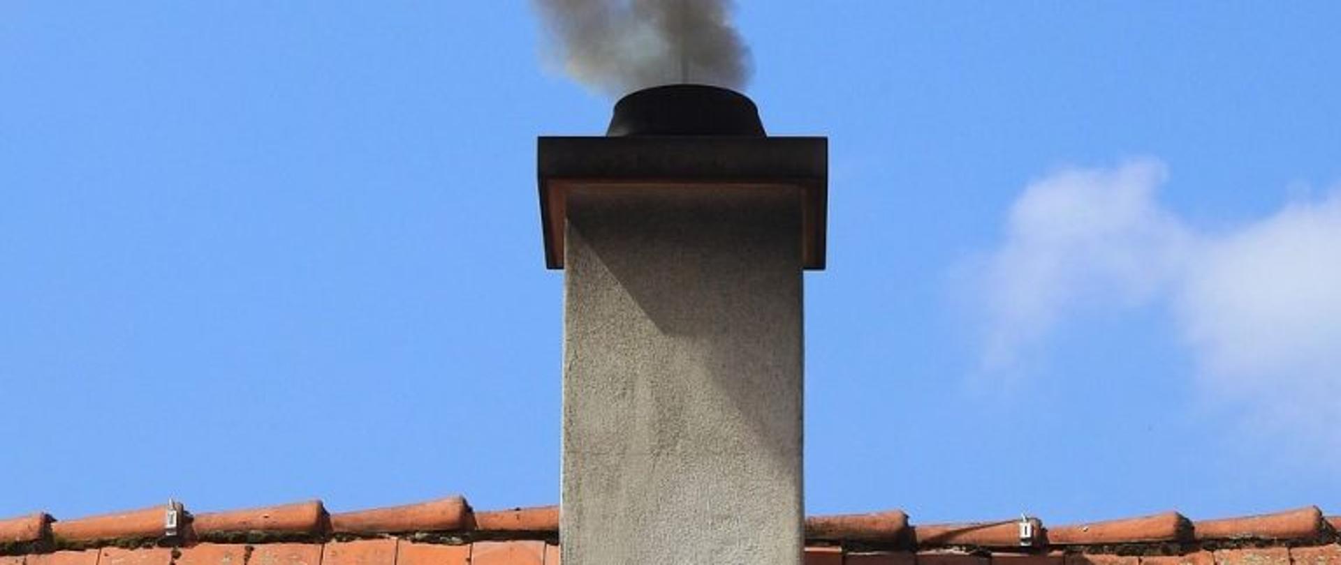 Na zdjęciu dymiący komin z kawałkiem dachu pokrytym dachówką ceramiczną koloru pomarańczowego.