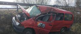 Widać czerwony samochód po dachowaniu w przydrożnym rowie, uszkodzony dach, otwarte drzwi od kierowcy i maska