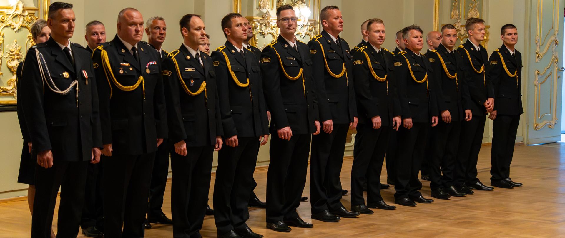 Bliskie ujęcie rzędu strażaków stojących na baczność podczas ceremonii. Mężczyźni są ubrani w ciemne mundury wyjściowe Państwowej Straży Pożarnej, ozdobione złotymi sznurami galowymi na prawym ramieniu. Wszyscy patrzą skupieni przed siebie. W tle widoczne są gładkie ściany sali z ozdobnymi, pozłacanymi elementami architektonicznymi