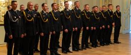 Bliskie ujęcie rzędu strażaków stojących na baczność podczas ceremonii. Mężczyźni są ubrani w ciemne mundury wyjściowe Państwowej Straży Pożarnej, ozdobione złotymi sznurami galowymi na prawym ramieniu. Wszyscy patrzą skupieni przed siebie. W tle widoczne są gładkie ściany sali z ozdobnymi, pozłacanymi elementami architektonicznymi