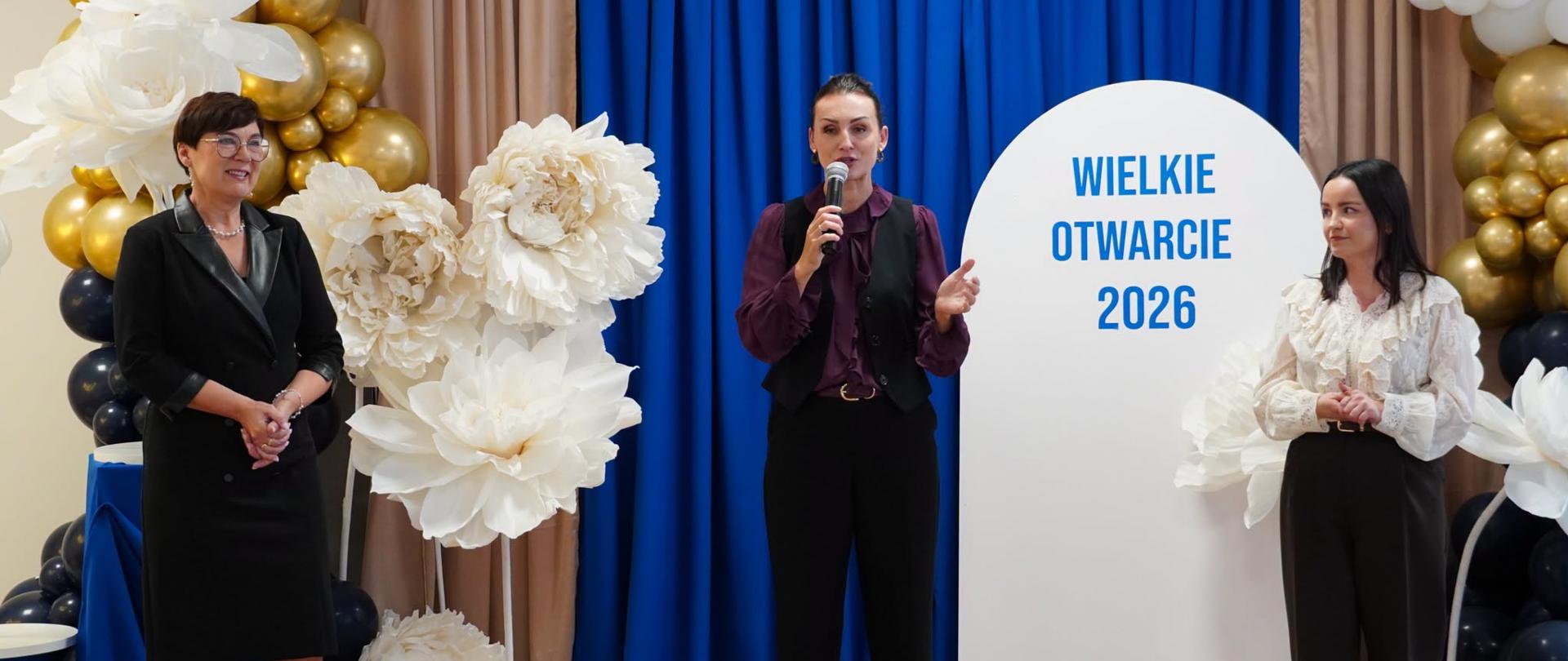 Na zdjęciu widoczne są trzy kobiety stojące na tle dekoracji przygotowanej z okazji wydarzenia. Kobieta stojąca w centralnej części, ubrana w fioletową bluzę, czarną kamizelkę i spodnie, trzyma w dłoni mikrofon i przemawia do zgromadzonych. Po jej lewej stronie stoi kobieta w czarnej sukience z jasnym kołnierzem, a po prawej kobieta w jasnej bluzce z falbanami oraz czarnych spodniach. W tle znajduje się niebieska kurtyna, kompozycja z białych i złotych balonów oraz sztuczne kwiaty, a także biała tablica z napisem „WIELKIE OTWARCIE 2026”.
