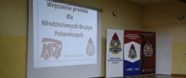 Napis na ekranie wręczenie promes dla Młodzieżowych Drużyn Pożarniczych obok stoją banery jednostek organizacyjnych Państwowej Straży Pożarnej. 