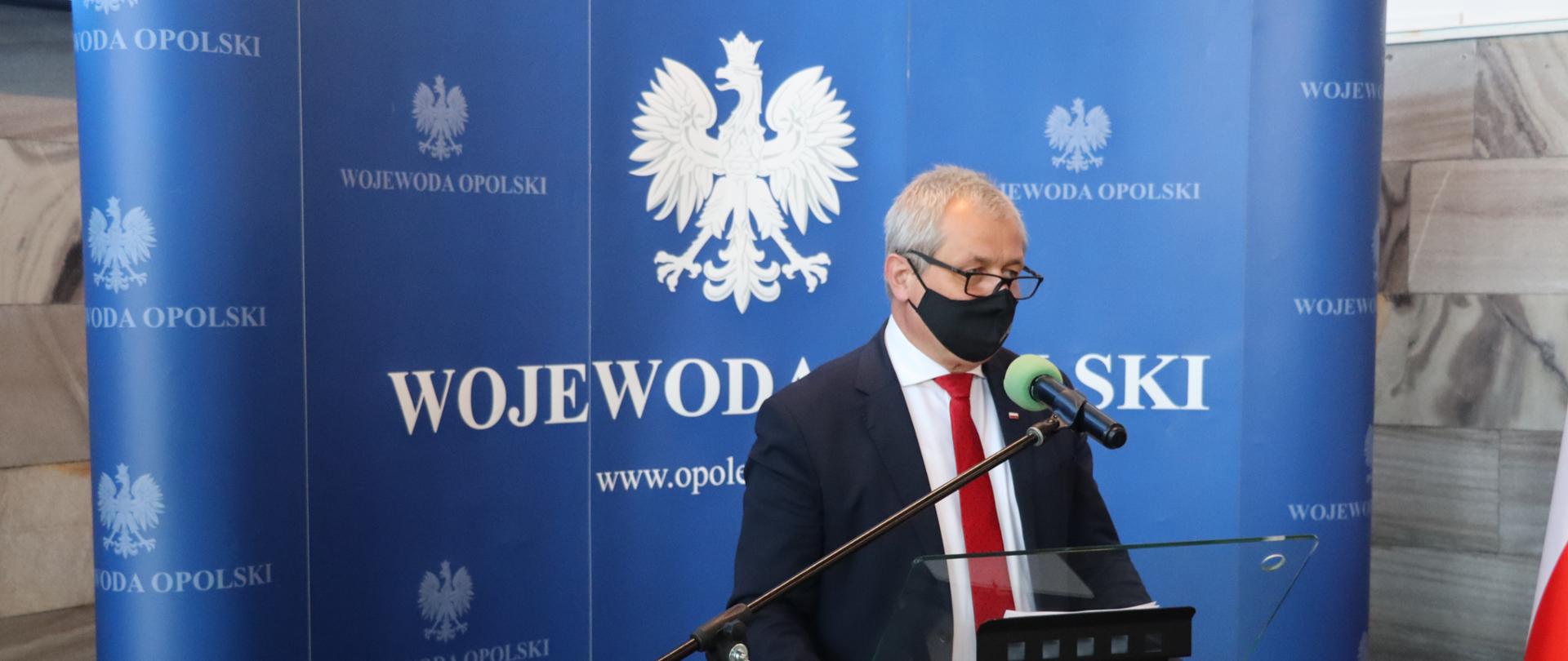 Na zdjęciu na środku kadru wojewoda opolski, stojący za szklanym pulpitem. W tle ścianka granatowa z napisem wojewoda i godłem. Po prawej stornie kadru widać flagę biało-czerwoną. Wojewoda przemawia do dziennikarzy, rękoma obejmuje szklany pulpit. 