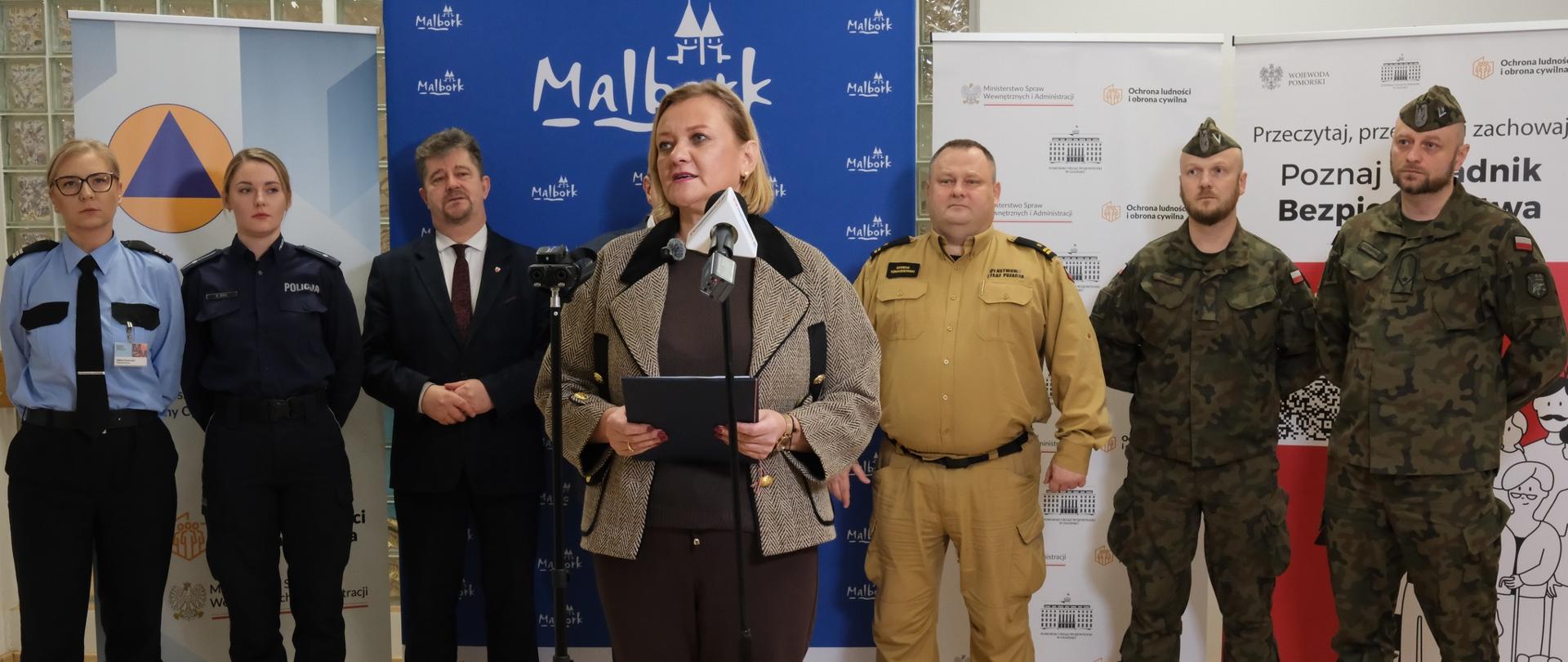 kobieta (wicewojewoda) przemawia przed mikrofonem, za nią kilka osób w tle baner z napisem Malbork
