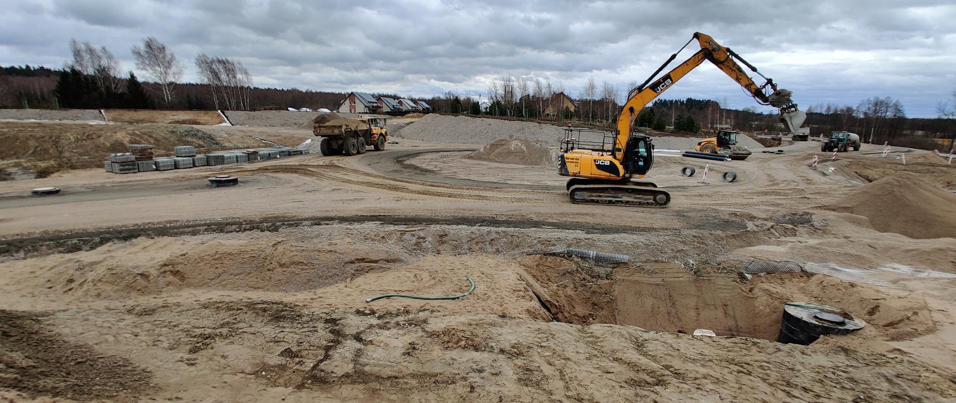 Roboty ziemne na placu budowy. Widoczne hałdy żwiru i maszyny budowlane. W oddali zabudowa jednorodzinna.