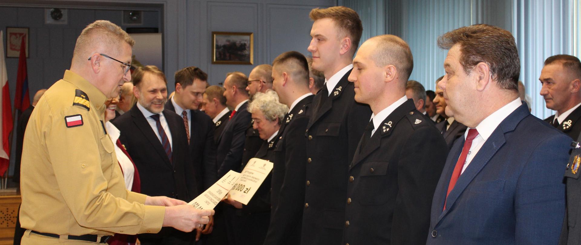 Zastępca KG PSP wręcza promesy , przed nim w rzędzie ustawieni są druhowie i przedstawiciele samorządu