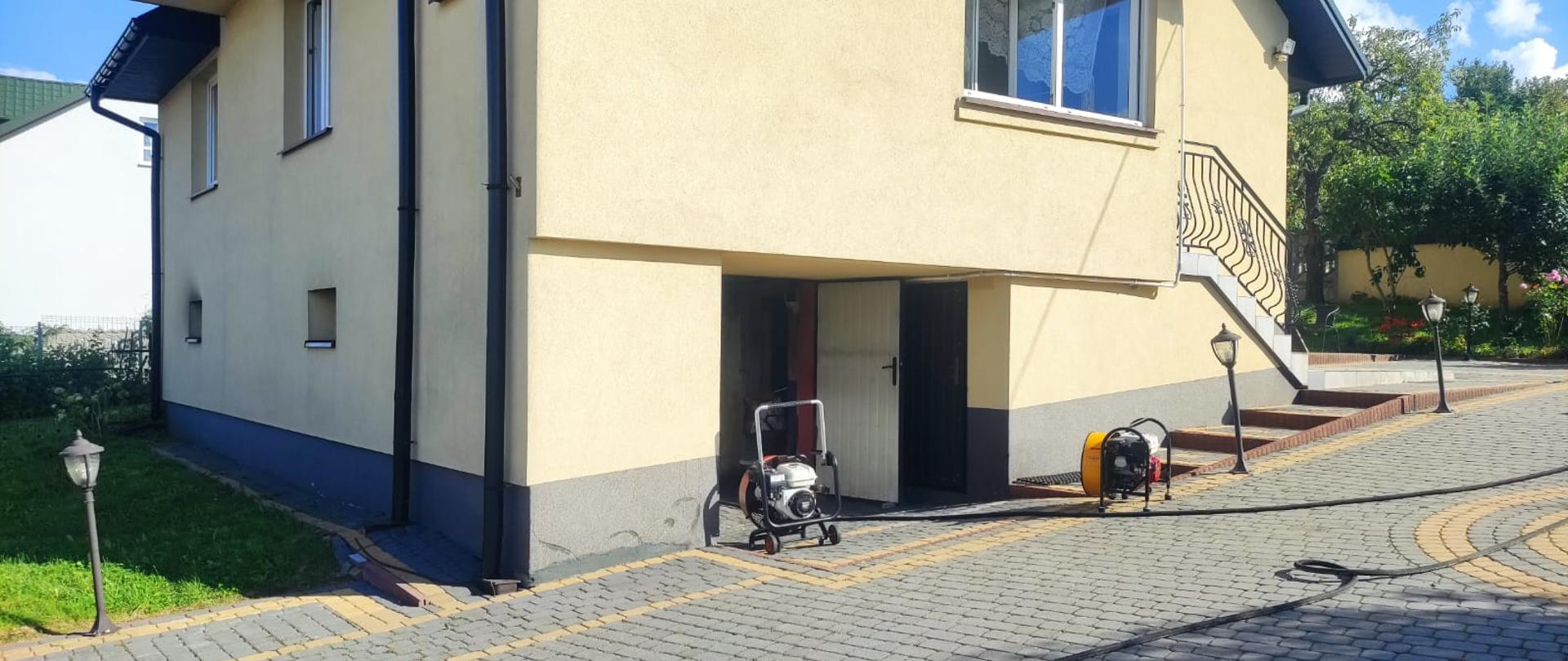 Na zdjęciu przedstawiono budynek jednorodzinnym, w którym doszło do pożaru. Drzwi do garażu są otwarte, a w ich świetle stoją dwa wentylatory, które wypychają dym z wnętrza przewietrzając pomieszczenia.