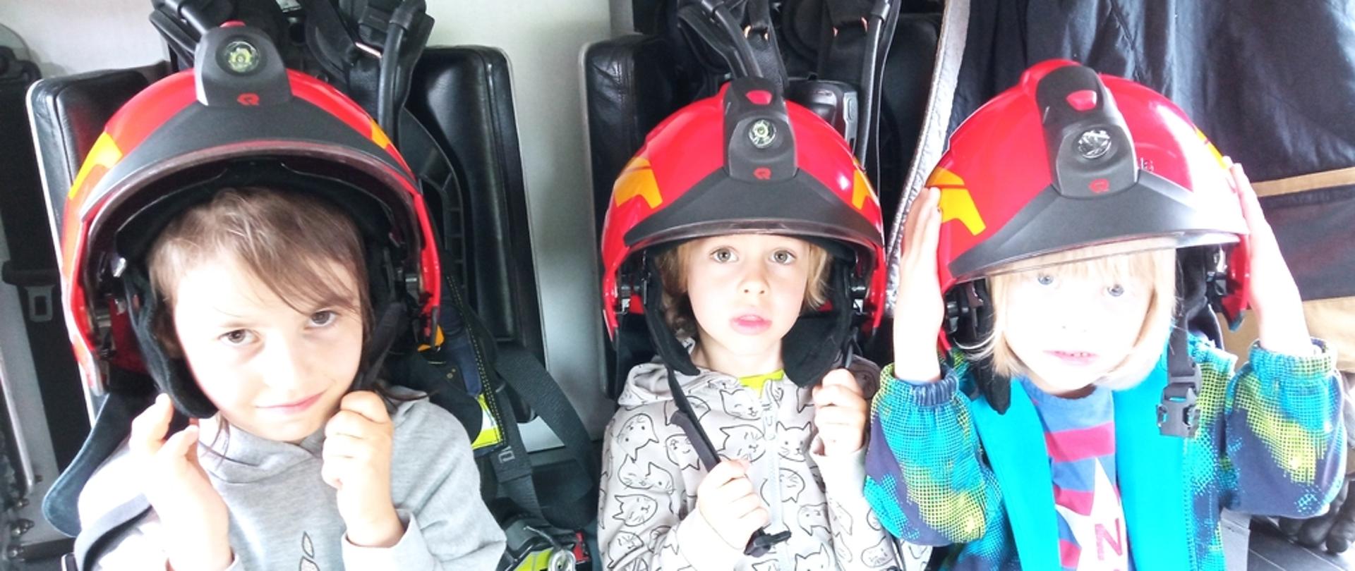 Na zdjęciu widoczne trzy dziewczynki w czerwonych hełmach strażackich siedzące w samochodzie pożarniczym.