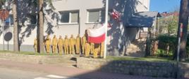 Grupa strażaków w jednolitych, żółtych mundurach stoi w szeregu przy ścianie jasnego budynku. Na pierwszym planie dwie osoby trzymają rozpostartą biało-czerwoną flagę, którą wciągają na maszt. Wokół widać drzewa rzucające cienie, chodnik oraz przejście dla pieszych, a całość to oficjalna ceremonia na świeżym powietrzu.