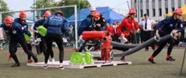 siedmioosobowa drużyna żeńska podczas bojówki pożarniczej, zawodniczki rozbiegają się z odcinkami wężowymi w rękach, na środku murawy stadionu stoi na drewnianym podeście motopompa do podawania wody, w tle widoczni zawodnicy oraz sędziowie