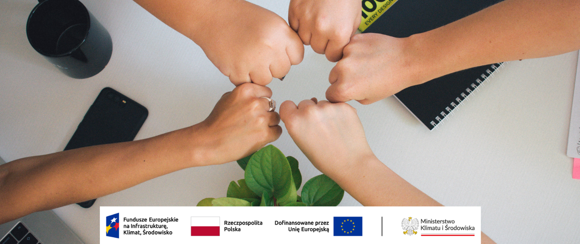 Pięć zbliżonych do siebie wokoło, zaciśniętych w pięść dłoni symbolizujących współpracę. W tle blat biurka oraz przybory jak kubek, telefon, notes, kwiat. Na dole logotypy unijne.