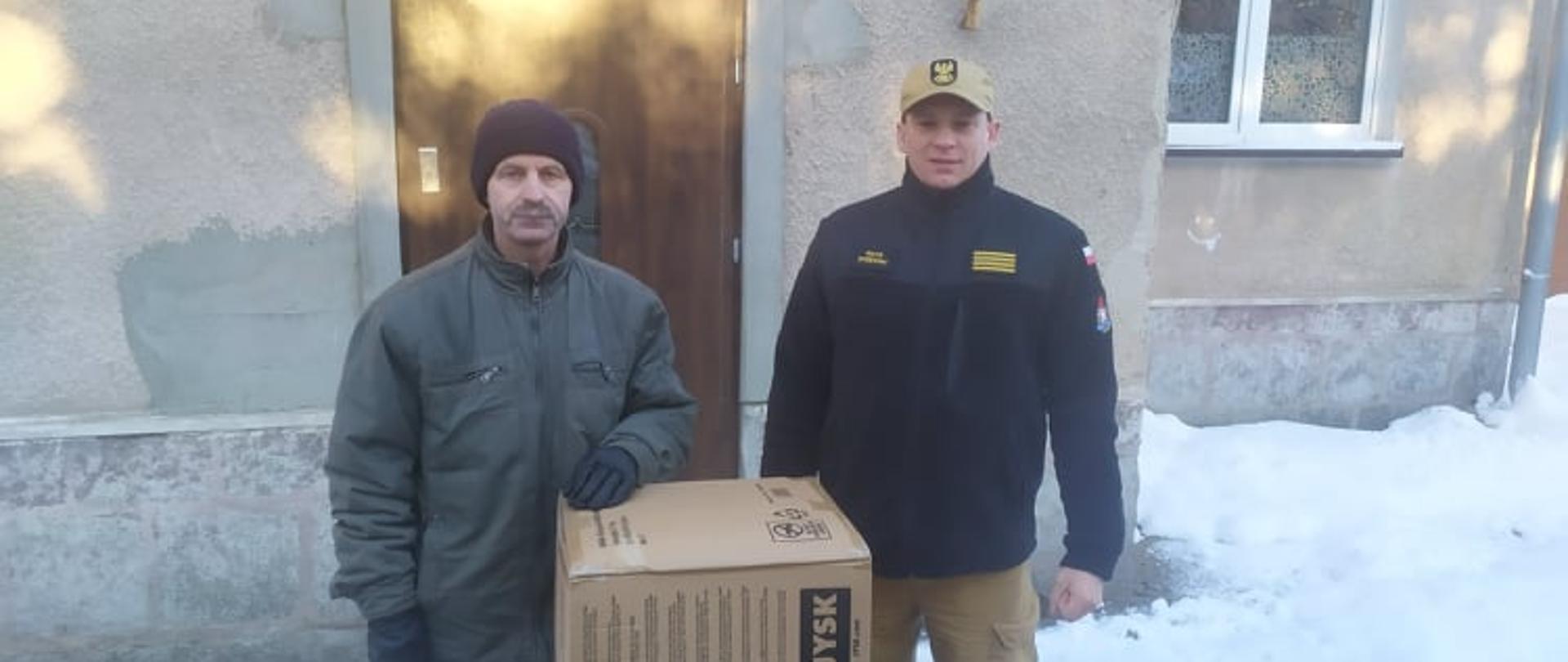 Pora dzienna, na zdjęciu stoi po lewej stronie właściciel spalonego domu w Monesterzyskach Wielkich ubrany w zimową kurtkę i czarną wełnianą czapkę. W środku na ziemi stoi duży karton do wysokości łokcia. Po prawej stronie stoi funkcjonariusz w czarnym polarze i jasnobrązowej czapce z daszkiem. W tle widać budynek.