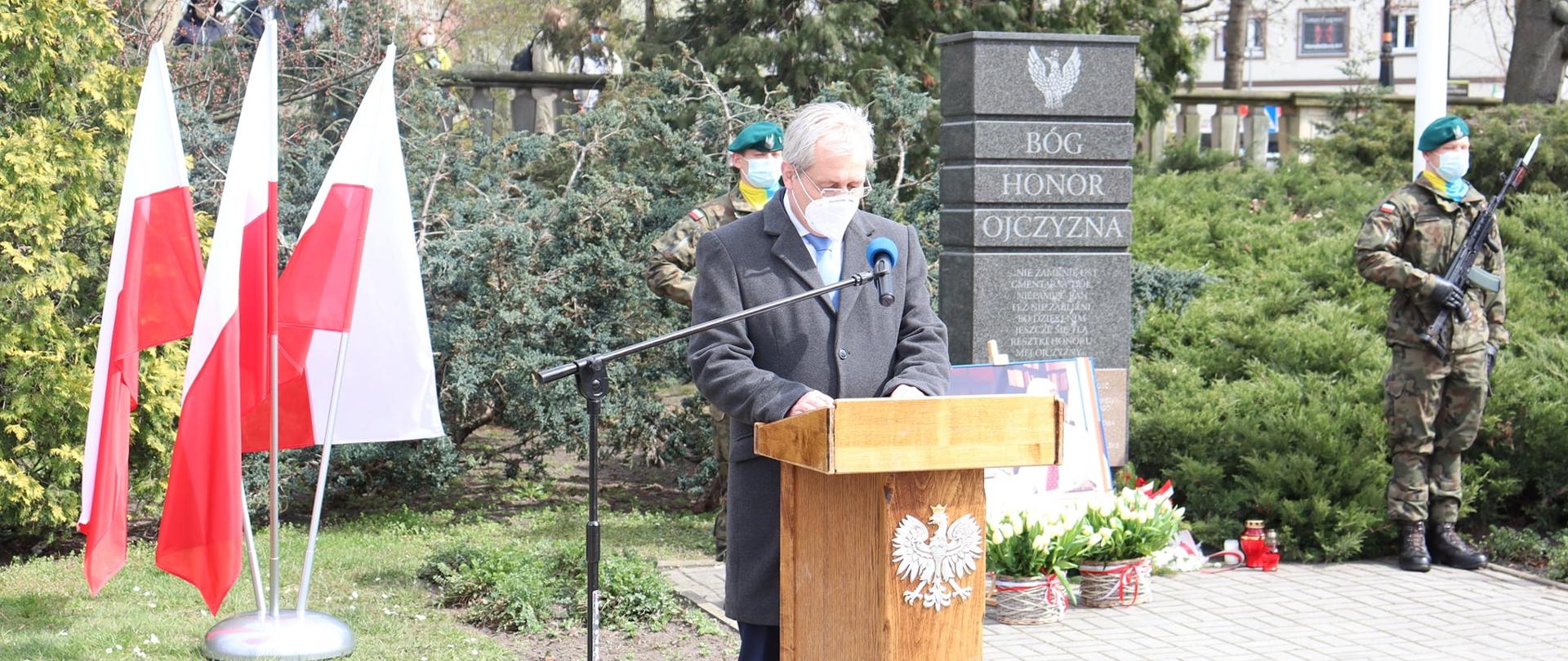 Na zdj. za mównica drewnianą z orzełkiem (w centralnej części kadru) stoi ubrany w granatowy płaszcz wojewoda opolski. Mężczyzna spogląda w dół - najprawdopodobniej na kartkę przemówieniem. W tle biało czerwone flagi, ustawione po obu stronach pomnika Żołnierzy Niezłomnych. Pomnik jest w czarnym kolorze - przypomina prostokąt. Na nim zapisano trzy wartości: Bóg, Honor, Ojczyzna. Przy pomniku wartę honorową pełnią żołnierze z 10 BLOG. Pod pomnikiem ustawione kwiaty, żółte tulipany. 