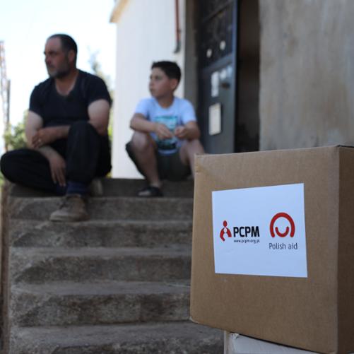 Na pierwszym planie po prawej stronie stoi paczka z logo Polskiej pomocy i PCPM, a w tle na schodach siedzi ojciec z synem