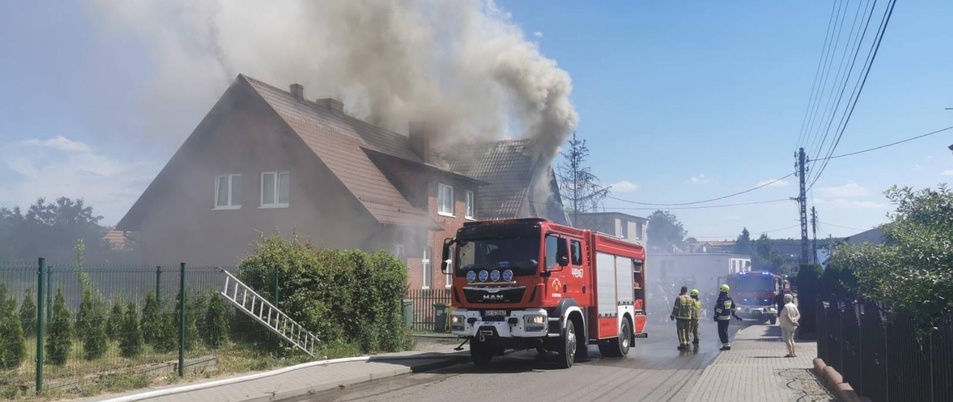 Unoszące się kłęby dymu nad dachem budynku objętego pożarem. Przed budynkiem na ulicy ustawiony samochód gaśniczy.