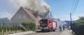 Unoszące się kłęby dymu nad dachem budynku objętego pożarem. Przed budynkiem na ulicy ustawiony samochód gaśniczy.