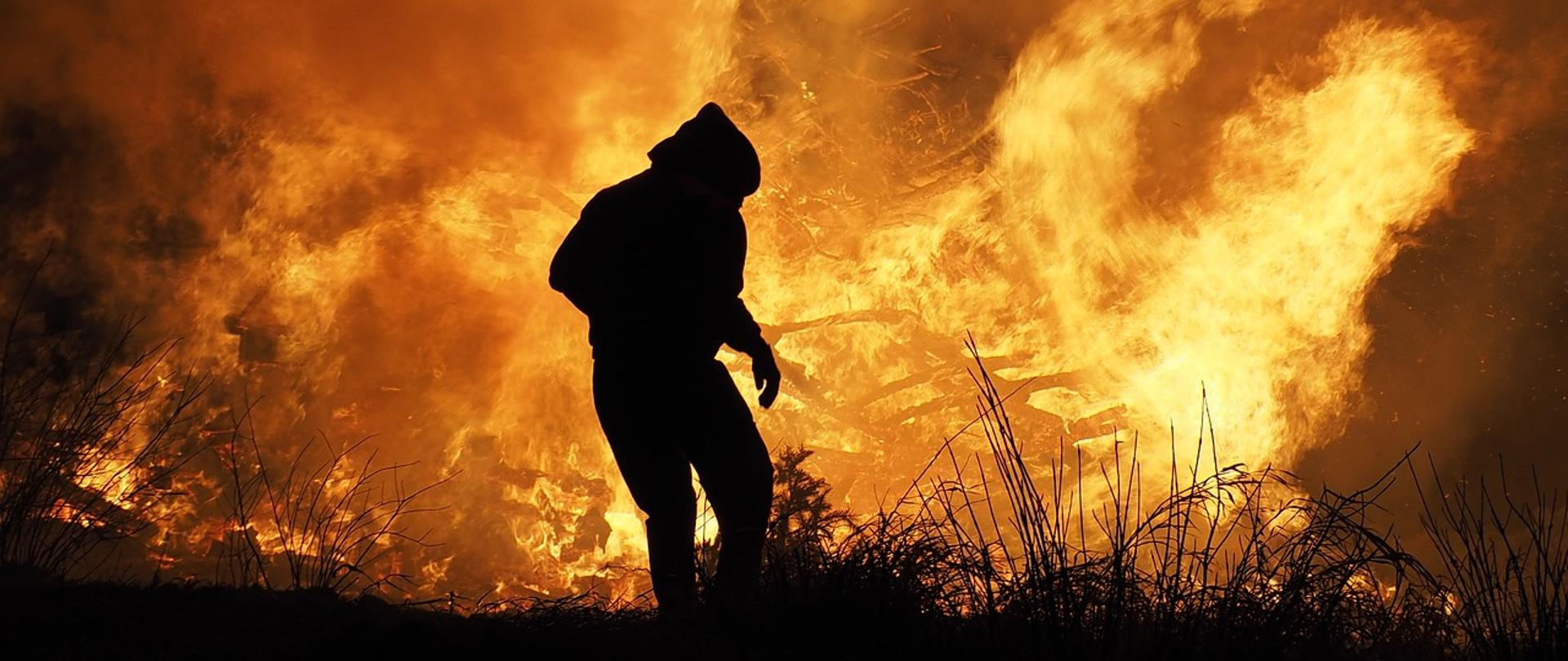 Zdjęcie przedstawia palące się trawy oraz zarys osoby na ich tle