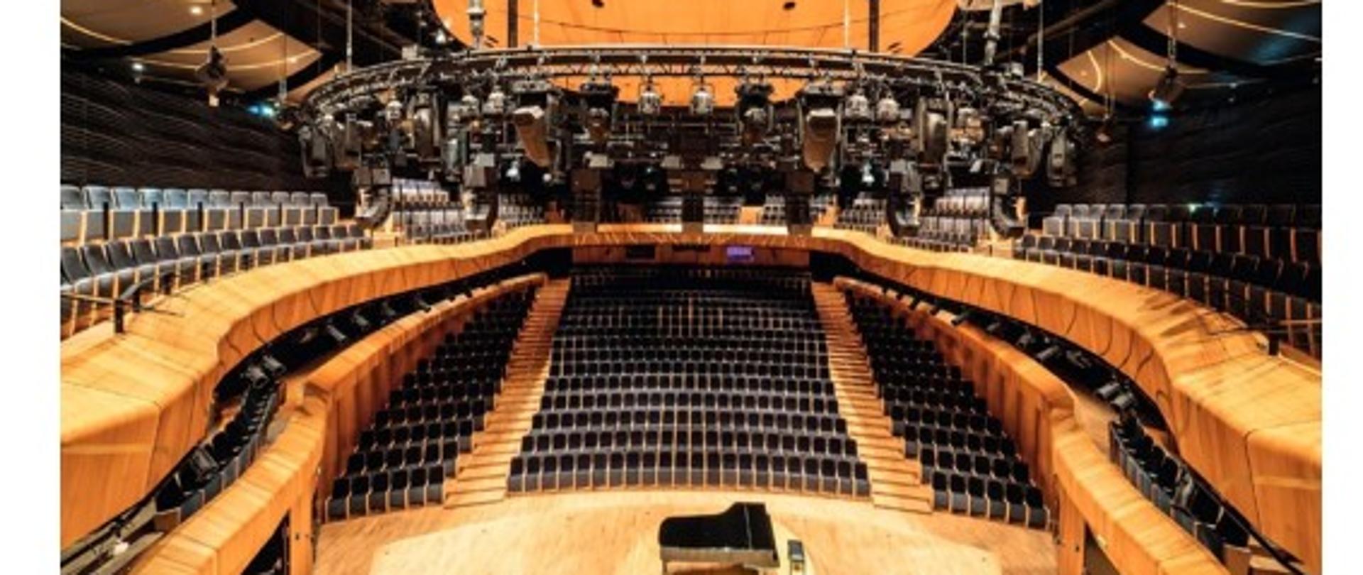 Zdjęcie składa się z dwóch części, przedstawiających zewnętrzny i wewnętrzny widok tej samej nowoczesnej budowli — Cavatina Hall.
Górna część zdjęcia: Przedstawia budynek z lotu ptaka, wykonany wieczorem lub w nocy. Budowla ma bardzo nowoczesny, futurystyczny kształt, przypominający nieregularną, organiczną formę. Na pierwszy plan wysuwa się duża, szklana kopuła w kolorze czerwonym, oświetlona od środka. Cały kompleks otoczony jest oświetlonym chodnikiem, a wokół widać ulice, samochody i okoliczne budynki. W tle widoczna jest zabudowa miejska, a atmosfera jest spokojna i wieczorna.
Dolna część zdjęcia: Pokazuje wnętrze sali koncertowej w środku budynku. Sala jest przestronna, z drewnianą sceną na środku, na której stoi fortepian koncertowy. Widownia otacza scenę półkoliście na kilku poziomach, tworząc elegancki i nowoczesny układ.
Nad sceną wisi duża konstrukcja oświetleniowa z wieloma reflektorami. Dominują ciepłe barwy drewna i ciemne, eleganckie fotele, co tworzy przytulną, a jednocześnie prestiżową atmosferę.