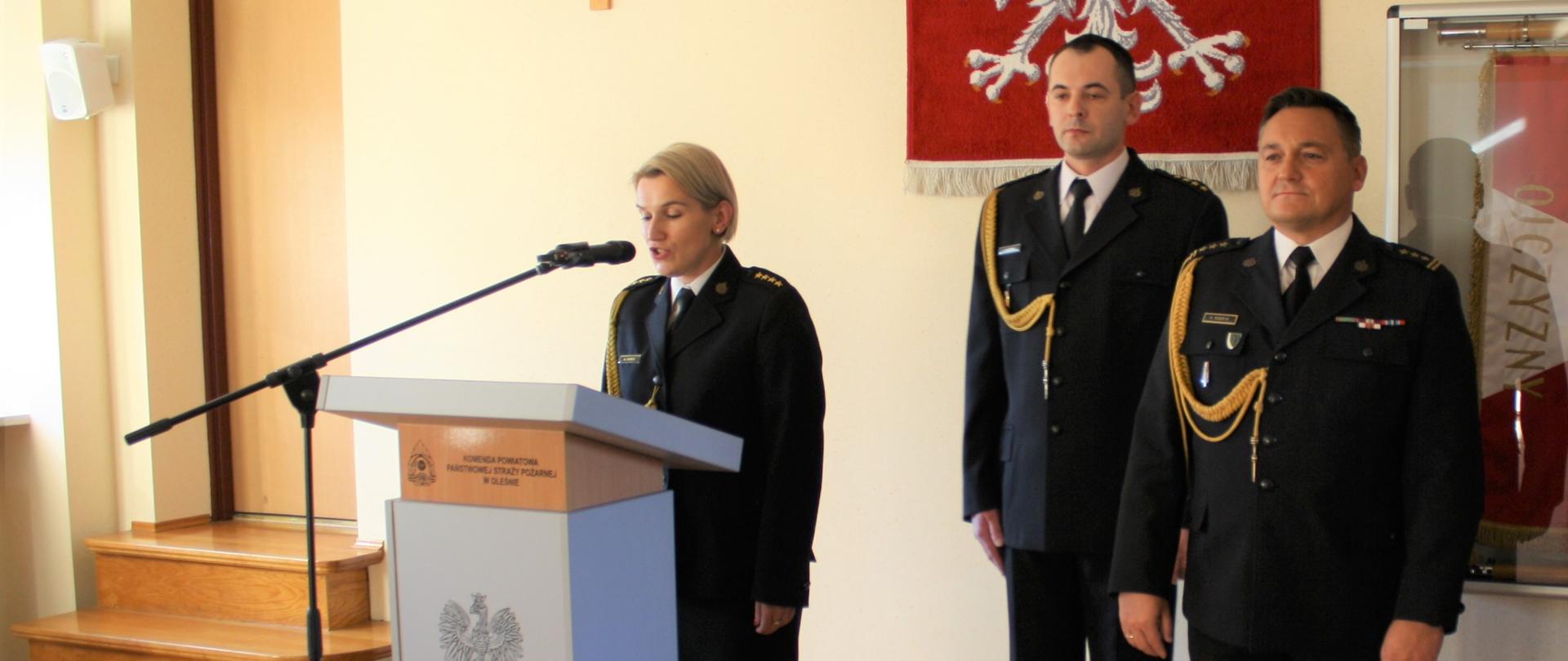 Na zdjęciu strażacy stoją w mundurach wyjściowych, jedna osoba przemawia za mównicą, na ścianie wisi godło Polski i Krzyż.