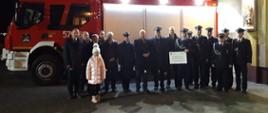 Wręczenie promesy w Pakosławiu. Plac przed remizą OSP w Pakosławiu. Pamiątkowe zdjęcie uczestników spotkania na tle wozu strażackiego. Po prawej stronie w gablocie oświetlona figura św. Floriana.