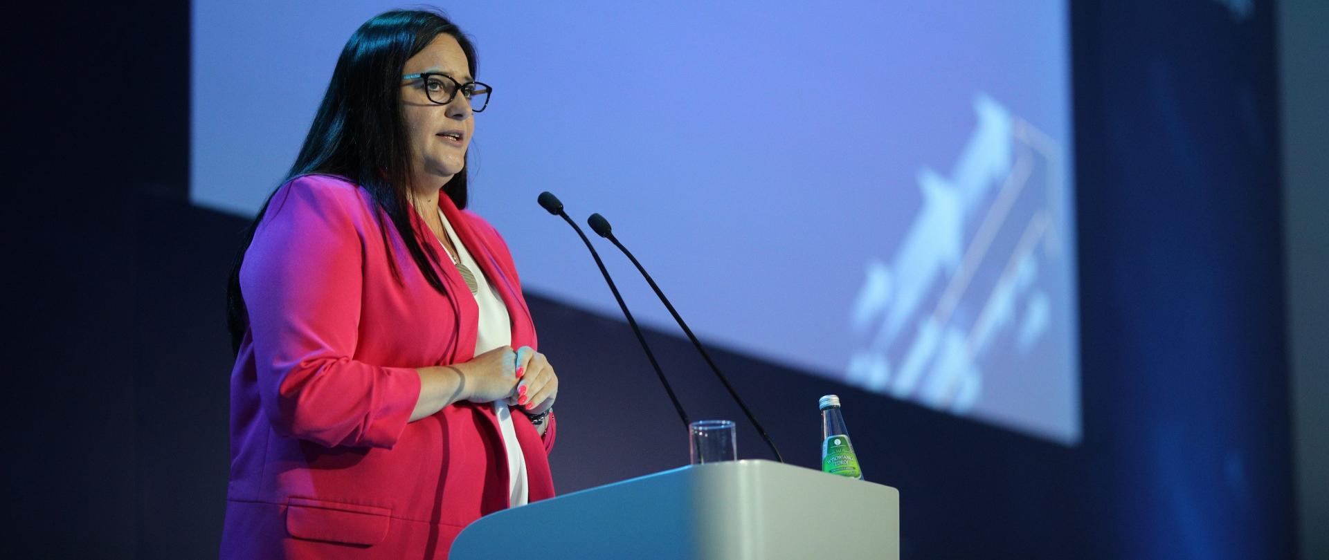 Minister Małgorzata Jarosińska-Jedynak stoi w mównicy. Za nia niebieski ekran.