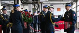 Moment wręczenia podziękowań. Nadbryg. Tomasz Baran w mundurze galowym przekazuje st. bryg. Ireneuszowi Szpyrce pamiątkową księgę. Obok nich funkcjonariuszka trzyma duży kosz czerwonych róż. Atmosfera jest serdeczna, strażacy uśmiechają się do siebie.