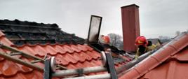 Zdjęcie dachu pokrytego blachodachówką. Jeden arkusz jest zdjęty, widać pod nim spalony dach. Otwarty jest wyłaz dachowy. Na dachy dwaj strażacy w czerwonych hełmach.