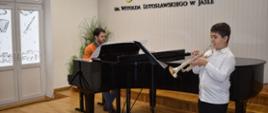 Na zdjęciu wykonanym w auli PSM widnieje uczeń grający na trąbce wraz z akompaniamentem fortepianu. Przy fortepianie nauczyciel Piotr Duda. Kolorystyka zdjęcia jest biało-czarno-brązowa. 