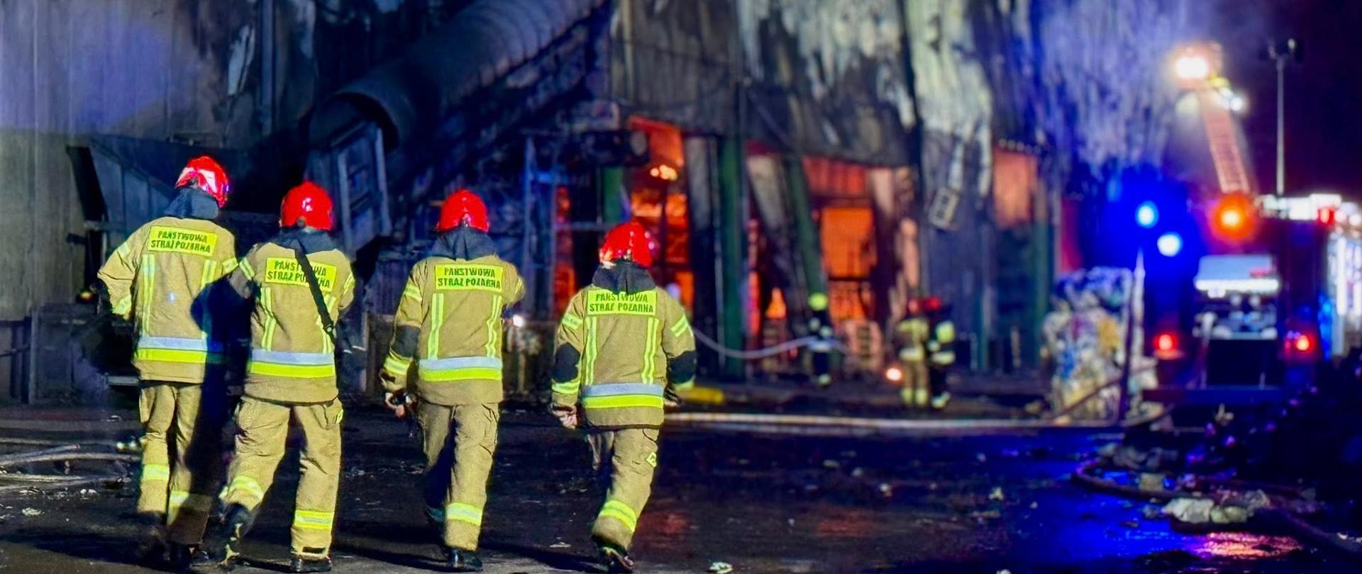 Sylwetki czterech strażaków zmierzających w kierunku palącej się hali. W tle widoczne płomienie. Obok hali pojazd pożarniczy z włączonymi niebieskimi światłami alarmowymi.