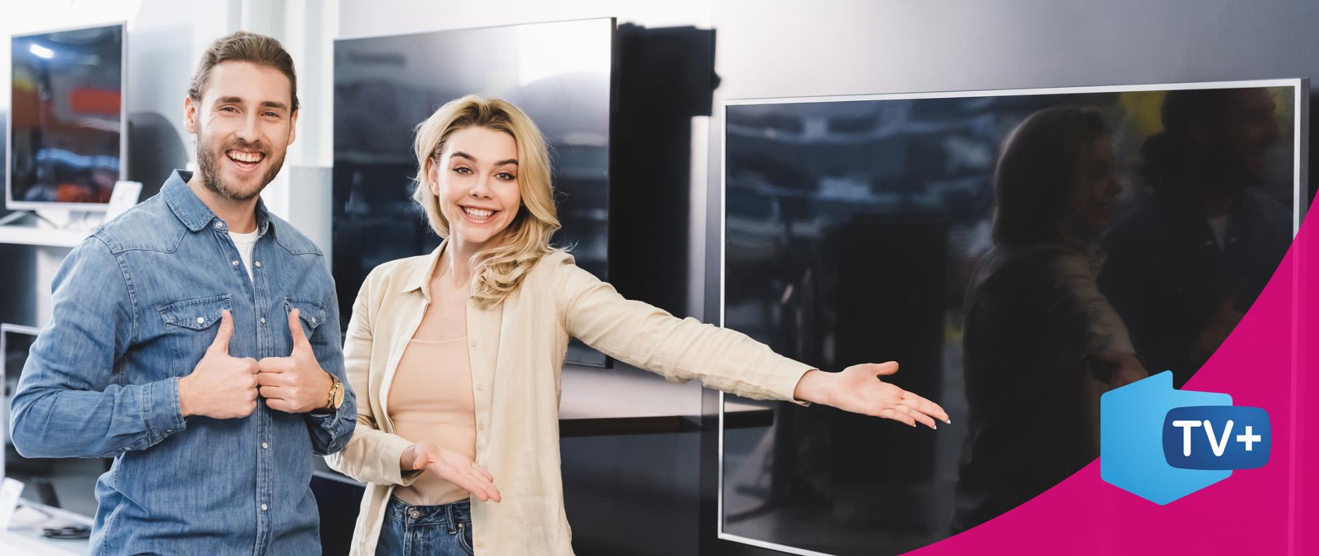 Na zdjęciu uśmiechnięta para młodych ludzi stojąca w sklepie RTV. Mężczyzna z zarostem, ubrany jest w jeansową koszulę - unosi w górę dwa kciuki. Stojąca obok kobieta, blondynka, ubrana jest w beżową koszulę i jeansy, wskazuje lewą ręką na nowoczesny telewizor. W tle widocznych jest też kilka innych telewizorów.