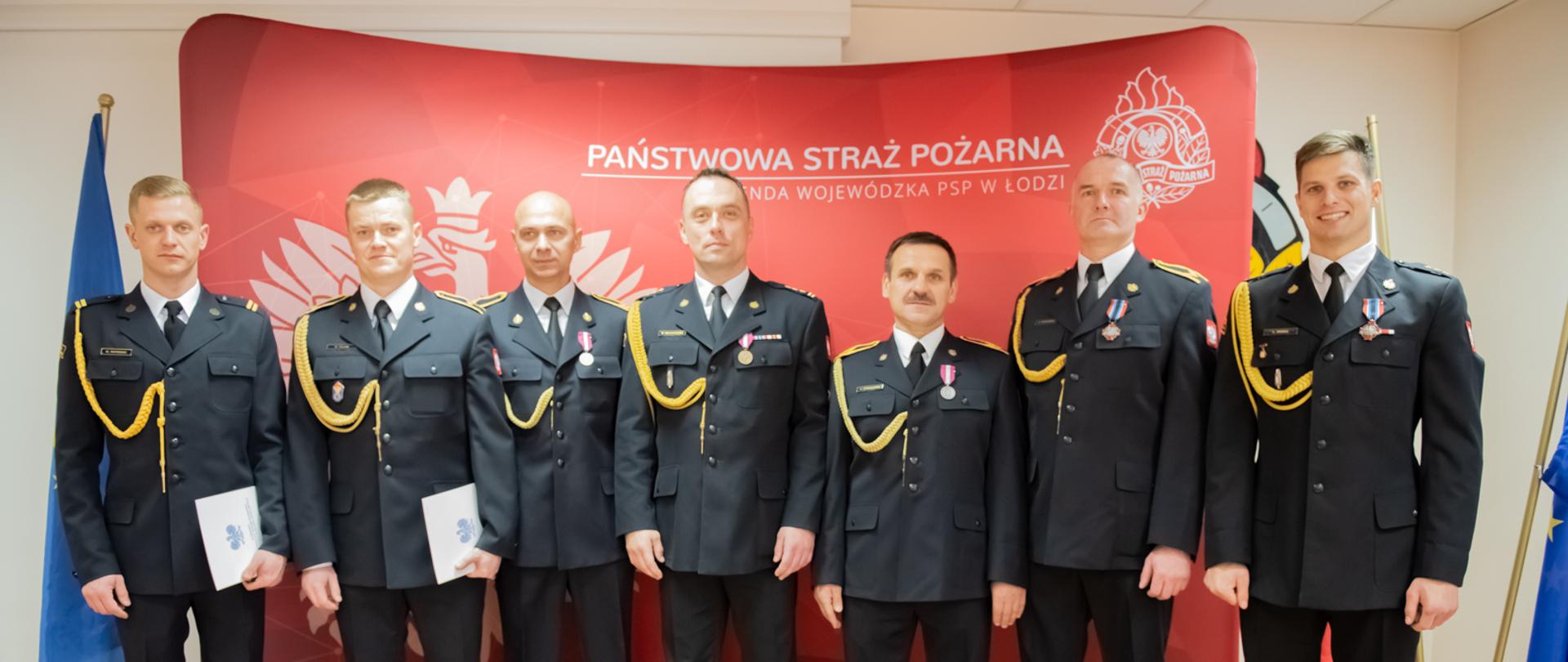 Na tle czerwonego baneru stoją w szeregu awansowani i odznaczeni strażacy.